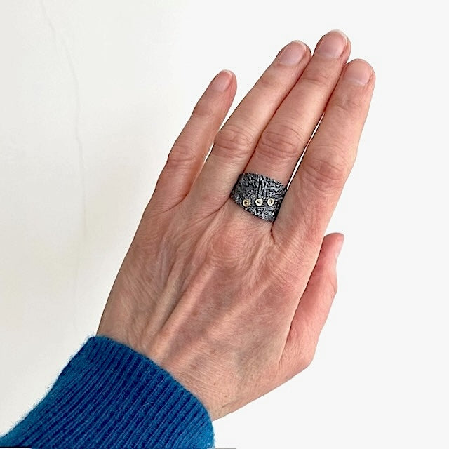 Cette bague en argent patiné et texturé se distingue par trois diamants taille brillant, sertis en or jaune sur dessus, alignés de côté. Bijou porté sur le majeur.