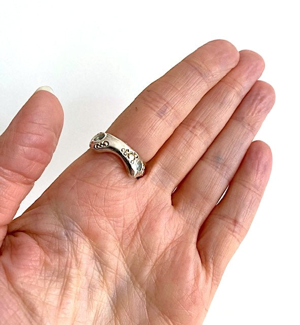 Bague en argent avec une aigue-marine en cabochon au centre et deux petites aigues-marines facettées sur l’anneau ciselé. La bague est portée à l’index, paume ouverte vers l’objectif.