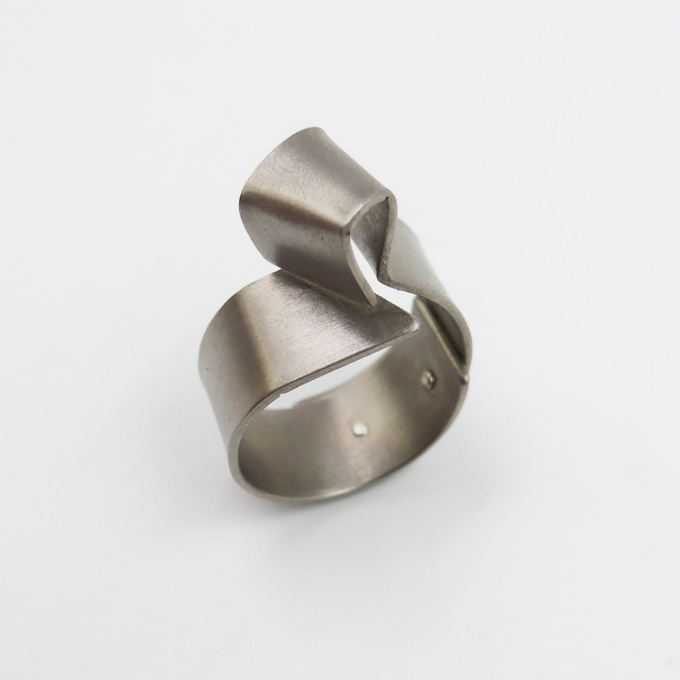 Bague sculpturale construite avec deux plaques de titane grises rivetées l'une à l'autre. Sur le dessus le titane est plié en forme de cube irrégulier. tandis que l'autre bande de métal s'enroule autour du doigt, comme un anneau large.