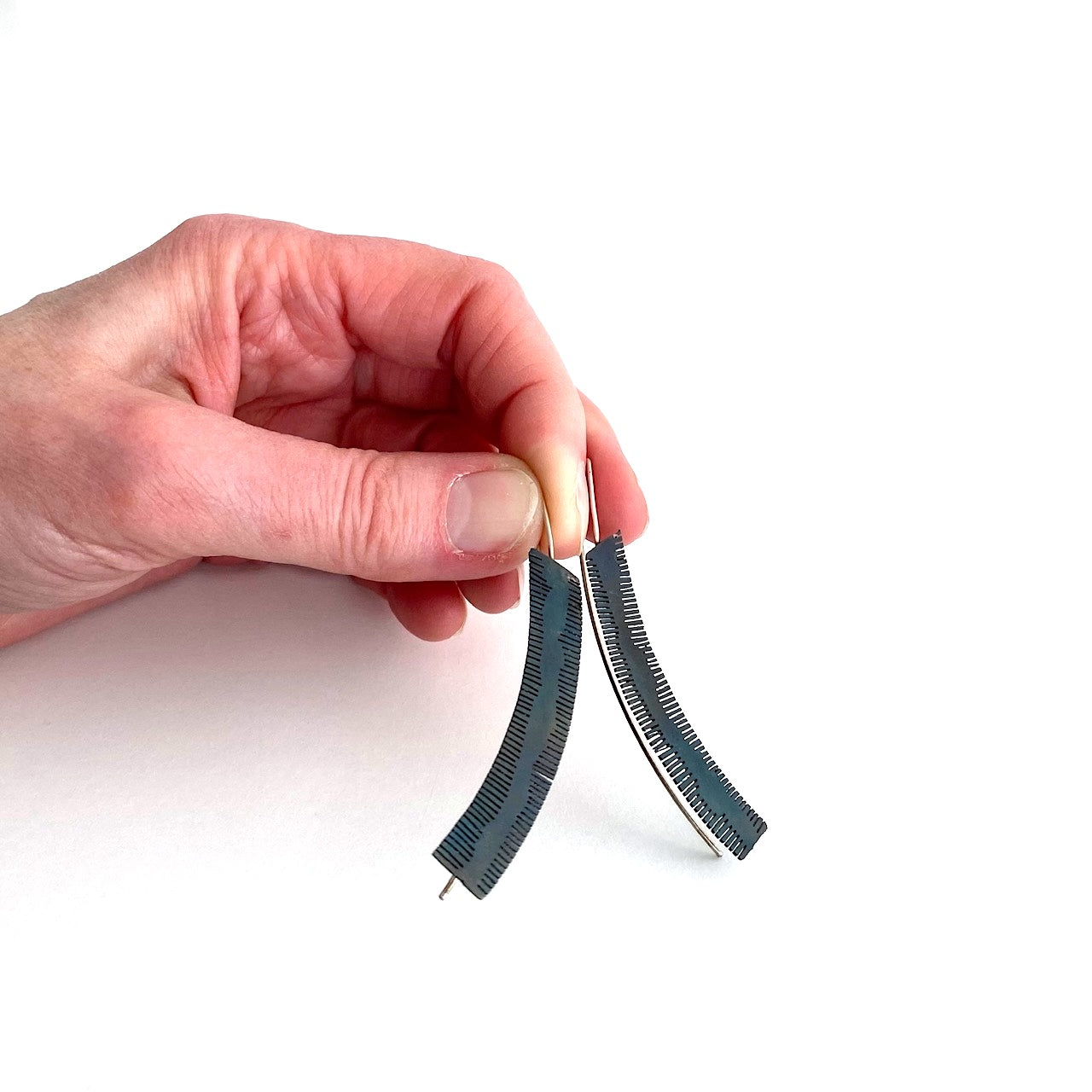 Boucles d'oreilles longues et géométriques découpées dans de l'acier patiné noir. Le métal scié sur la longueur du métal rappelle la délicatesse d'une plume. Une tige d'argent pour fermoir prolonge le bijou. Les boucles sont tenues par une main.