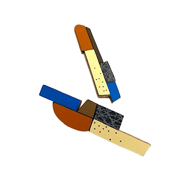 Des boucles d’oreilles longues et asymétriques associant des éléments géométriques en bois et formica. Ces pièces uniques, réalisées à la main par l’artiste belge Karen, mêlent des couleurs multiples : bleu foncé, marron chaud, jaune pâle, bronze métallisé, aux finitions mates ou avec motifs texturés.