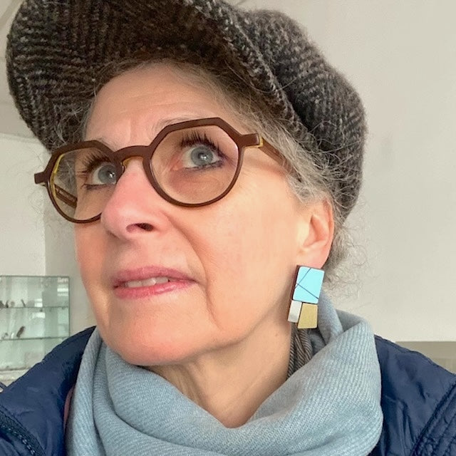 Boucles d’oreilles géométriques de forme rectangulaire en bois et formica bleu clair, gris, et doré métallisé texturé. Une création artisanal de l'artiste belge Karen Vanmol. Bijou porté vue de prfil.