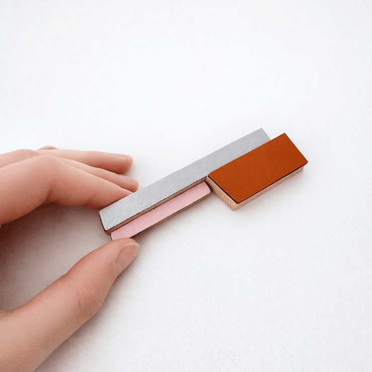 Broche contemporaine allongée en bois et formica gris,rose et brun orangé. Détail du bijou tenu entre deux doigts. Pièce unique réalisée à la main par l’artiste belge Karen.