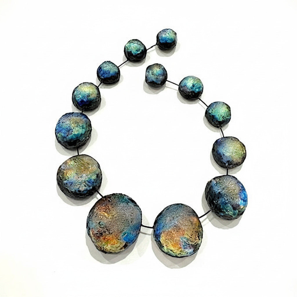 Collier sculpturale composé 13 sphères de tailles différentes en résine texturée et irisée, dans des nuances de noir bleuté, vert et reflets changeants. Les éléments sont assemblés sur un câble fin noir et résistant. Fermeture aimantée. Fabrication artisanale et française.