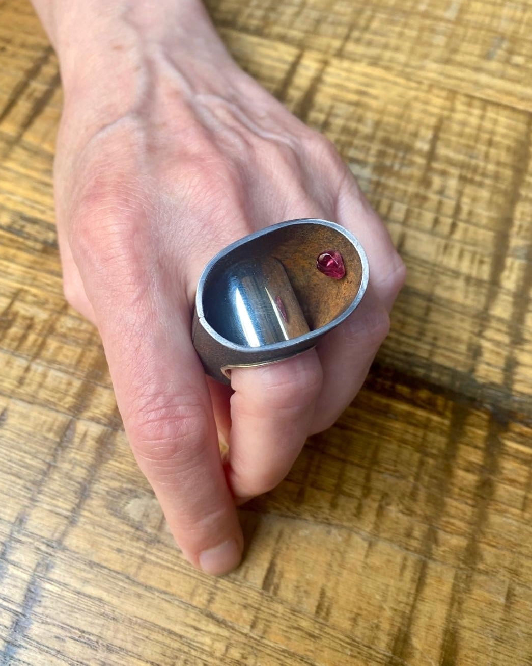 Bague haute et ovale en fer rouillé montée un large anneau douille en argent poli. À l'intérieur de la bague, sur le côté une tourmaline rose est cachée. La main est posée sur une table en bois.