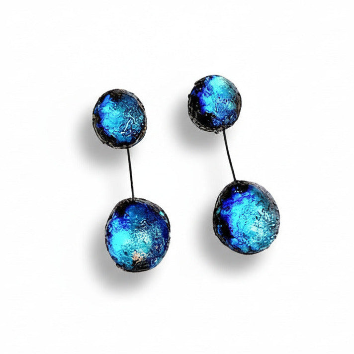 Boucles pendantes composées de deux sphères en résine irisée bleue, reliées ensemble par un câble noir fin et solide. Pour oreilles percées.
