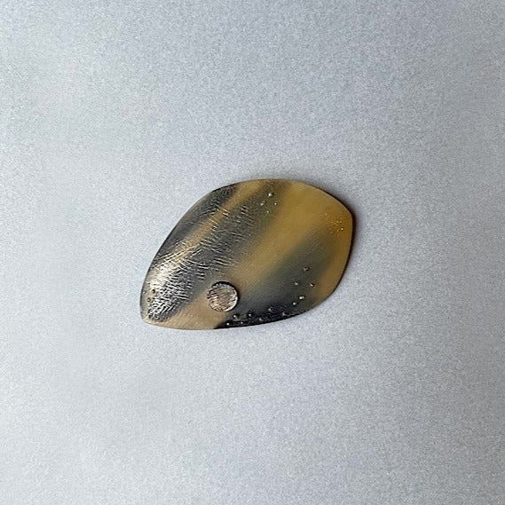 broche ovale, sculptée en corne dorée aux dessins bruns naturels. Le bijou est gravé, orné d'une pastille de nacre grise et de fil de nylon perlé.