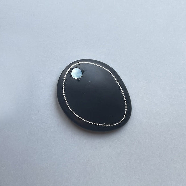 Cette broche ovale en corne noire est décorée d’une frise composée d'une rangée de petits points blancs peints. Une pastille de nacre grise apporte une touche d'éclat, au bijou.
