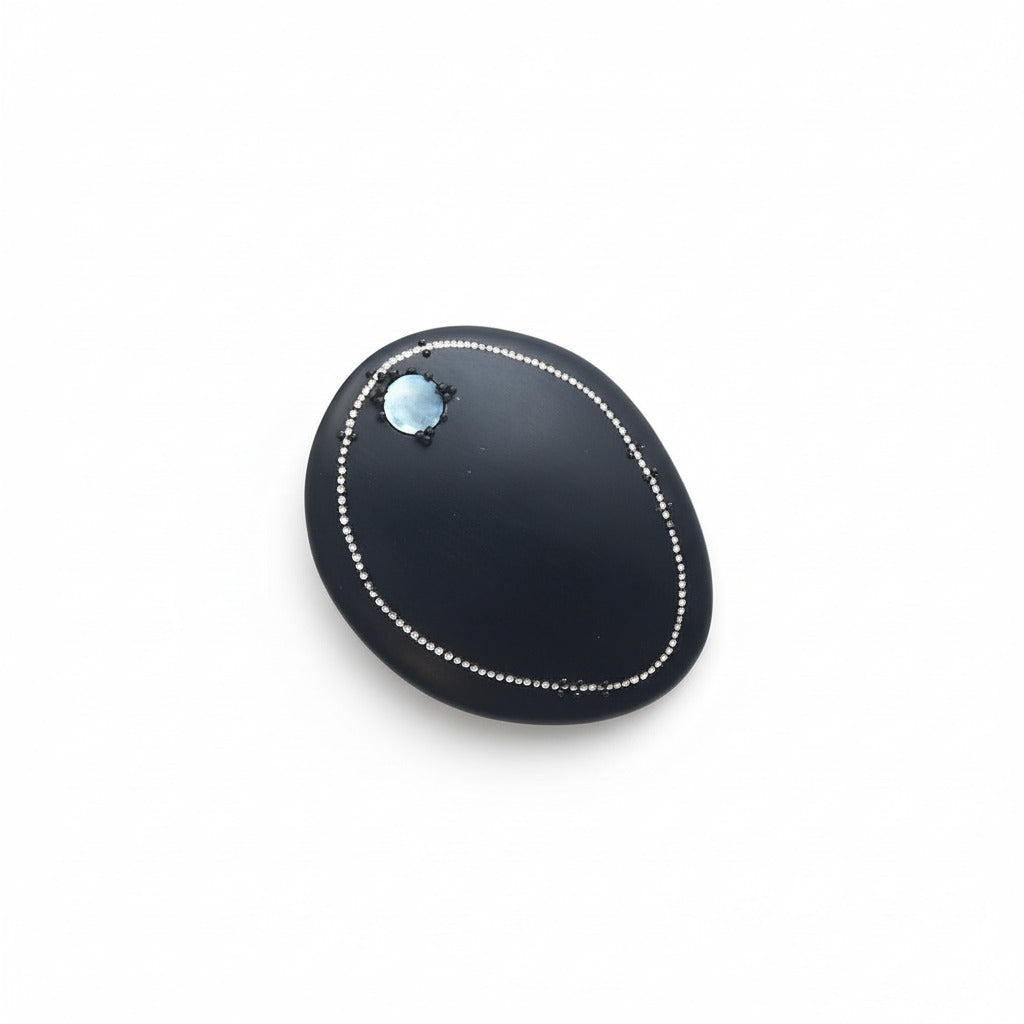 Cette broche ovale en corne noire est décorée d’une frise composée d'une rangée de petits points blancs peints. Une pastille de nacre grise apporte une touche d'éclat, au bijou.