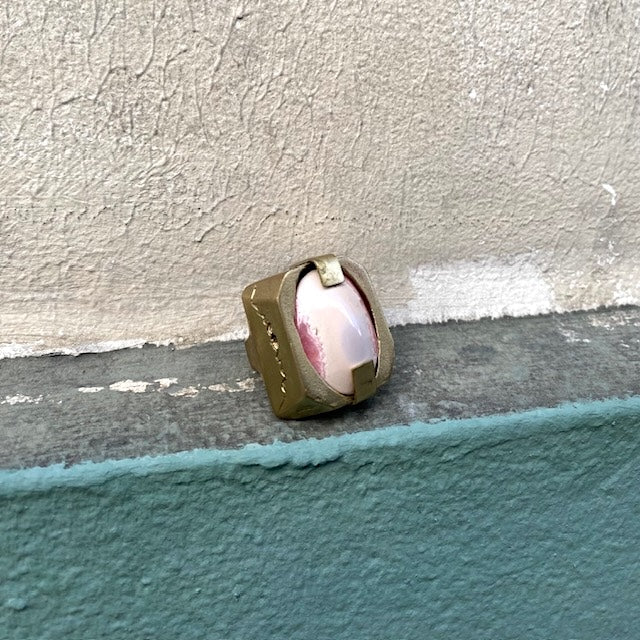 Bague sculpturale rectangulaire en bronze entaillé sur les longueurs. Au centre en cabochon, une Rhodochrosite ovale, rose tendre et fuchsia est tenue par deux griffes. Décor mur vert.