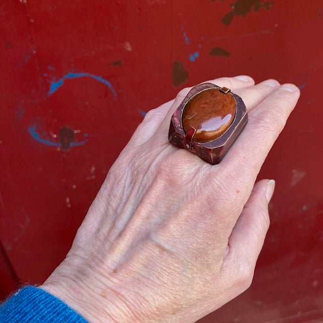 Bague sculpturale rectangulaire en bronze et peinture acrylique rouge, sur le dessus un jaspe rouge ovale est serti par 2 griffes de chaque côté. Bague portée, décor rouge.