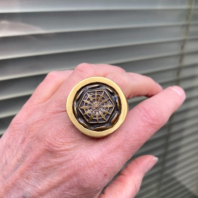 Large chevalière en bronze avec au centre un bouton ancien en fer décoré d'une rosace. Bague portée, décor store vert.