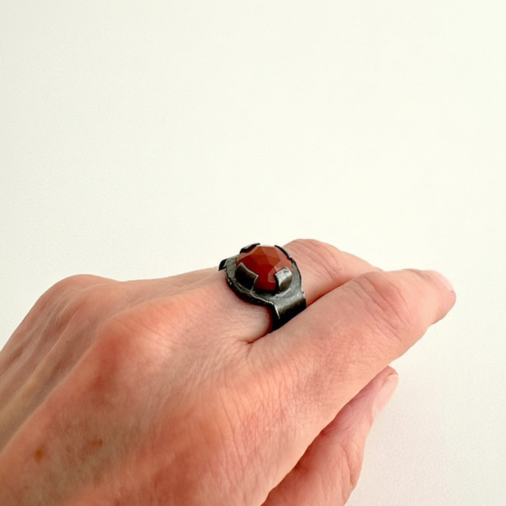 Chevalière contemporaine en argent oxydé avec sertissage quatre griffes plates sur jaspe rouge facetté. Bijou artisanal pensé comme une petite architecture portée. Détail, bijou porté.