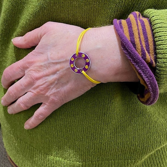 Bracelet Ready-Made composé d'une rondelle en aluminium violet sertie de six saphirs jaunes facettés. Bracelet design et unisexe présenté sur lien de coton ciré. Bijou porté.