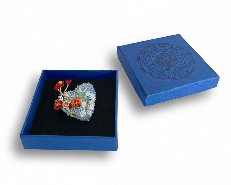 Prête à offrir, La broche Heart-Blossoms est présentée dans un élégant écrin bleu signé Trovelore, accompagnée de sa notice explicative détaillant le travail de broderie et le savoir-faire artisanal.