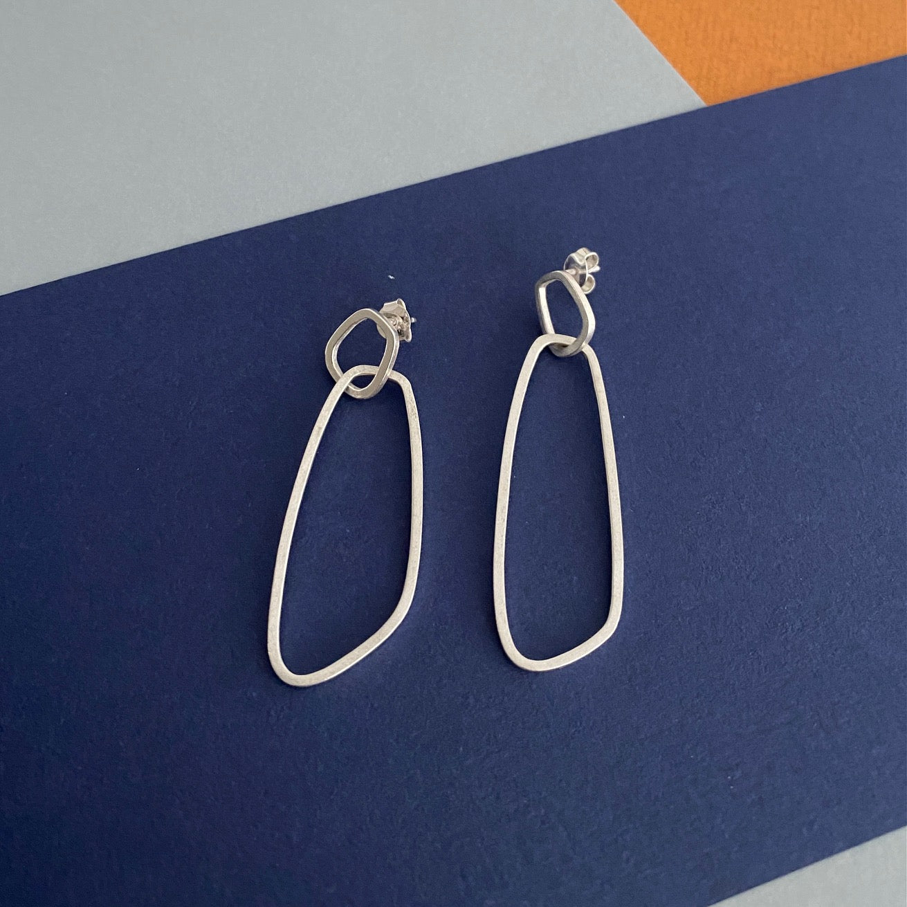 Boucles d’oreilles pendantes en argent satiné de type mobile composées de deux éléments posées géométriques sur fond couleurs bleu, gris et jaune.