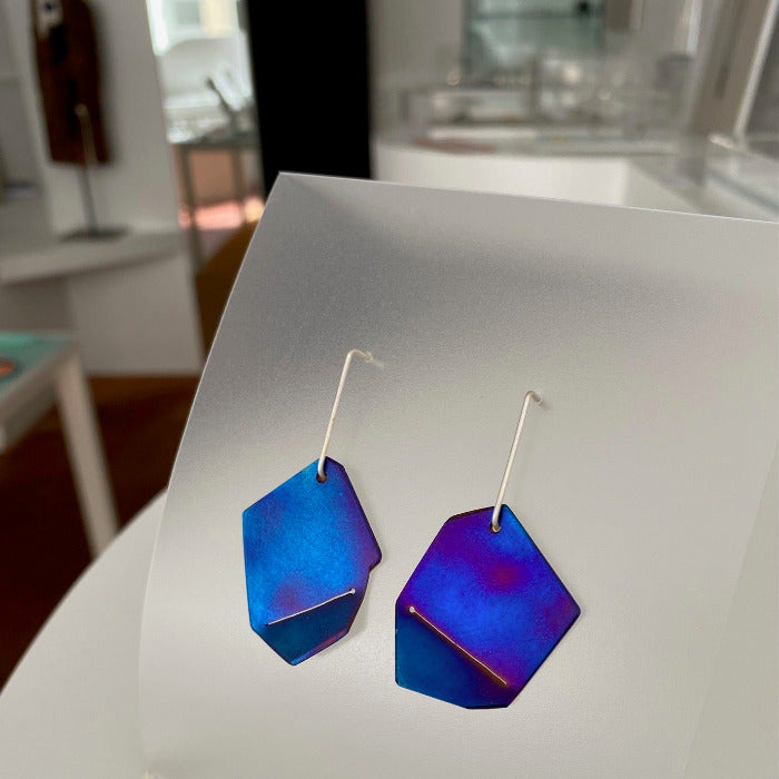 Grandes boucles d'oreilles titane plié et scié bleu foncé, tiges argent présentées fond décor galerie