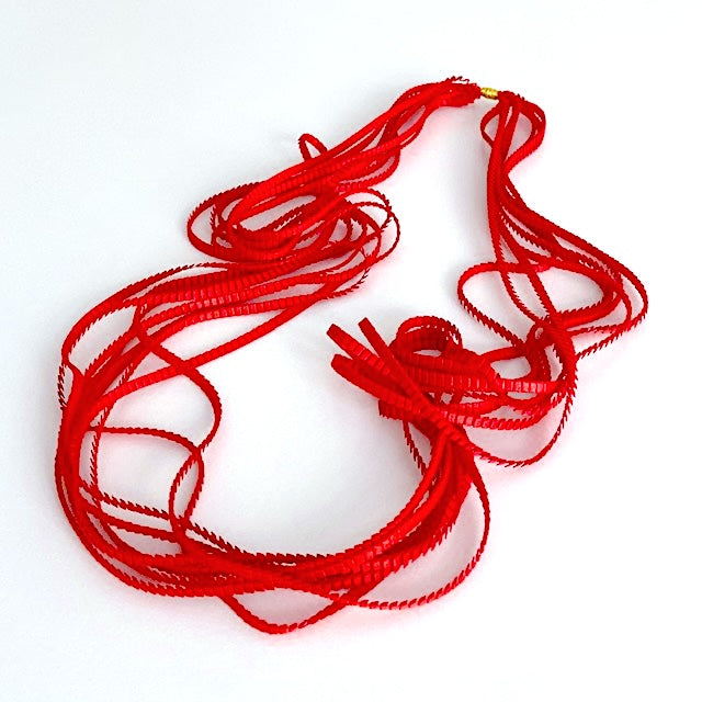 Long sautoir rouge composé de huit bandes de trois millimètres de satin plissé reliées les unes aux autres par un lien de coton jaune d'or.