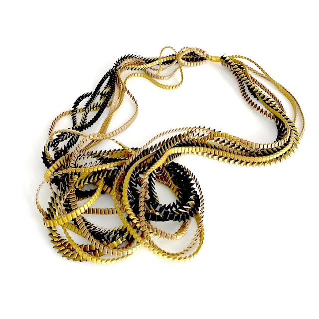 Long sautoir de couleur doré, olive et noir composé de huit bandes de trois millimètres de satin plissé reliées les unes aux autres par un lien de coton jaune d'or. 