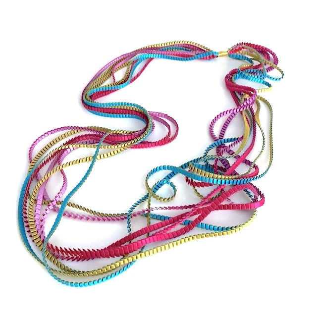 Long sautoir composé de huit bandes de trois millimètres de satin plissé de couleur, fuchsia, doré, turquoise, lilas,reliées les unes aux autres par un lien de coton jaune d'or. 