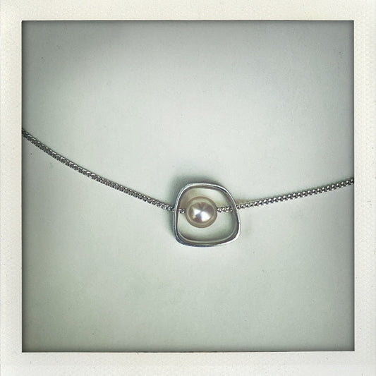 Collier en argent avec pendentif géométrique et perle d’eau douce banche. Fabrication artisanale en argent texturé traversé par une chaîne, création Anne Milbeau.
