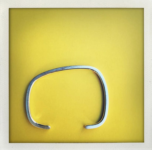 Bracelet géométrique en argent forgé au fil carré, jonc ouvert au design asymétrique. Modèle mixte, surfaces striées et intérieur poli. Bijou design contemporain, bijou minimaliste présenté sur un fond jaune.