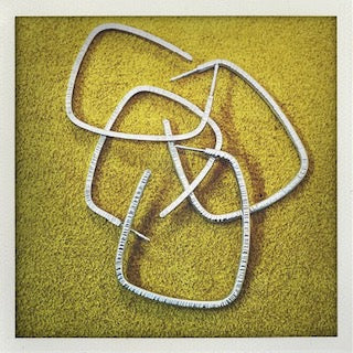 Détails, grandes boucles créoles géométriques en argent forgé en fil carré  strié posées présentées sur fond couleur jaune. Créations artisanales Anne Milbeau.