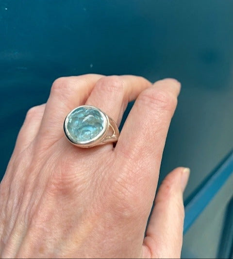 Bague argent ornée d'une aigue-marine en cabochon . La pierre sculptée dissimule une opale révélant des reflets changeant. L'anneau en argent ciselé présente deux aigues-marines facettées.