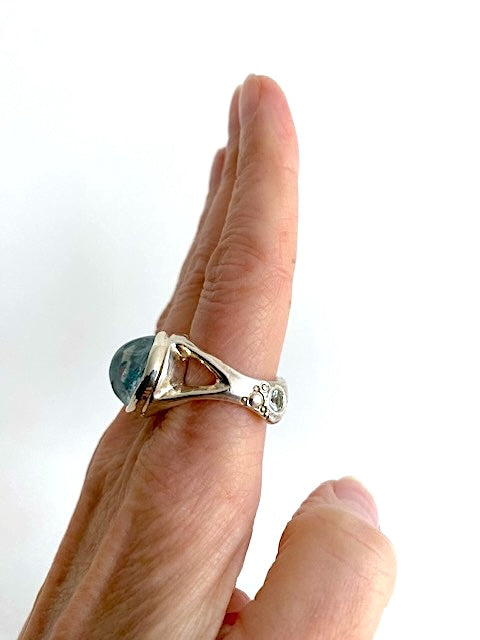 Bague en argent avec une aigue-marine en cabochon au centre et deux petites aigues-marines facettées sur l’anneau ciselé. La bague est portée à l’index, main de profil vers l’objectif.