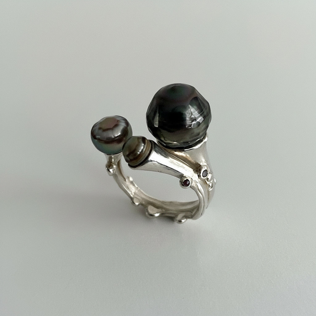 Cette bague ouverte, façonnée en argent ciselé, met en scène trois perles singulières : deux perles de Tahiti sculptées aux reflets changeants et une perle keshi. Une bague contemporaine, élégante et délicate réalisée à la main en pièce unique par Ivan Michelson, créateur français.