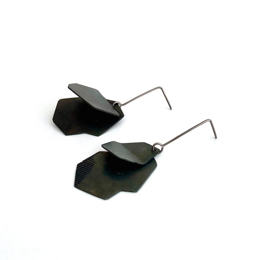 Boucles pendantes architecturales en acier patiné noir, plié et scié par endroit en lamelles. Elles sont montées sur des tiges droites en titane.