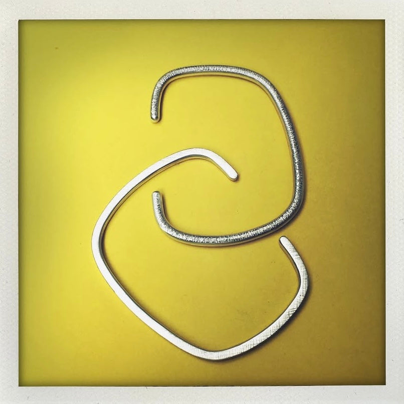 Deux bracelets contemporains, l'un des jonc ouvert est fabriqué en argent a fil carré et l'autre en fil rond,finition argent texturé et martelé. Présentés sur fond jaune. Créations géométriques signées Anne Milbeau.