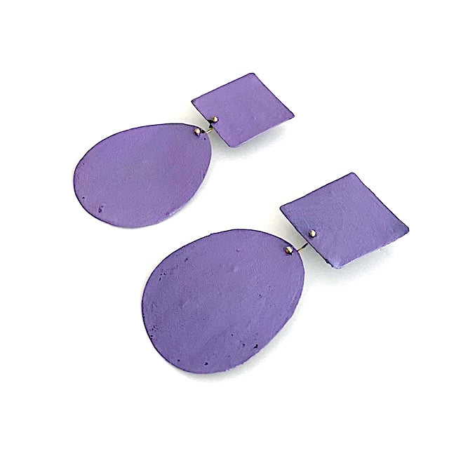 Ces boucles d'oreilles présentent un élément ovale suspendu à un élément carré placé sur l’oreille, composant une silhouette géométrique contemporaine.La surface est peinte à la main avec des pigment acrylique violet. Vue de profil. 
