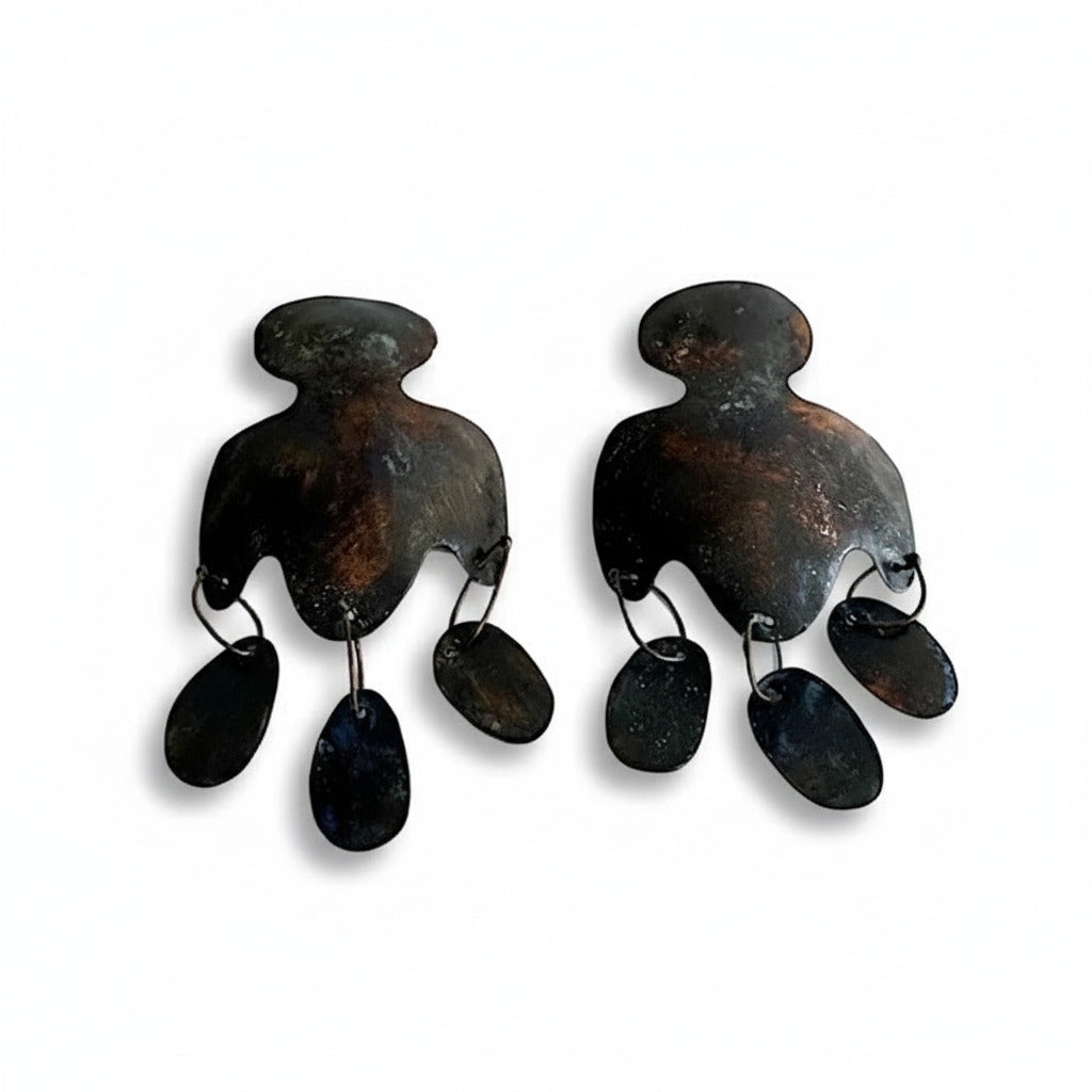 Cette paire en bronze laminé et pigments acryliques brun et noir se distingue par une découpe aux lignes arrondies et de trois pampilles allongées, alliant minimalisme et inspiration historique. Bijou façonné à la main par la créatrice greque Dora Haralembaki.