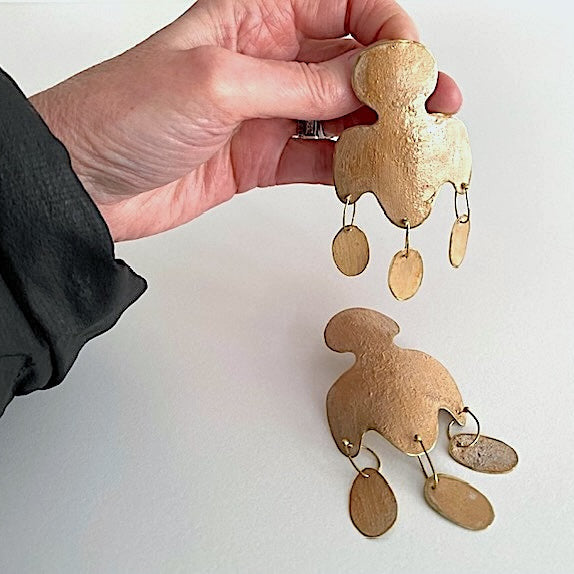 Détails d'une paire de boucles pendantes en bronze laminé et peint avec des pigments beige se distingue par une découpe aux lignes arrondies et de trois pampilles allongées. Une création artisanal de la créatrice grecque Dora Haralambaki.