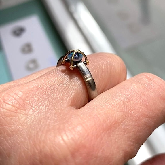 Une Opale en cabochon est présentée sur un anneau en acier inoxydable mat. Deux éléments d'or jaune entrecroisés viennent entrelacer la pierre. Bijou porté.