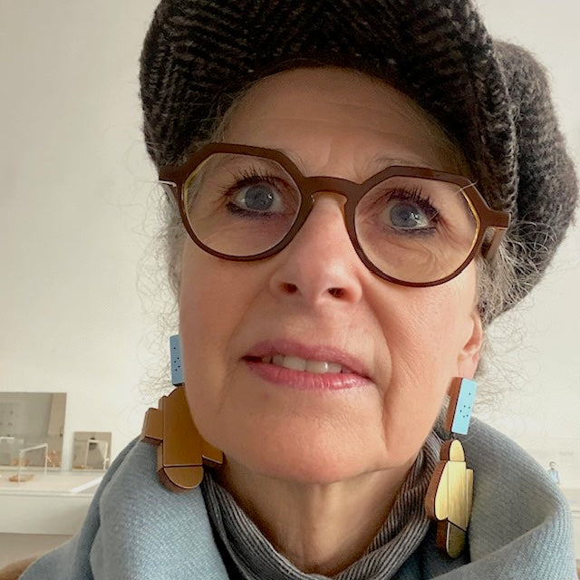 Suspendues à un support rectangulaire bleu clair à petits points noirs, ces boucles XL associent bois, formica doré texturé et doré métallisé. Légères et sculpturales, elles sont faites à la main par l’artiste belge Karen Vanmol. Bijoux portés.