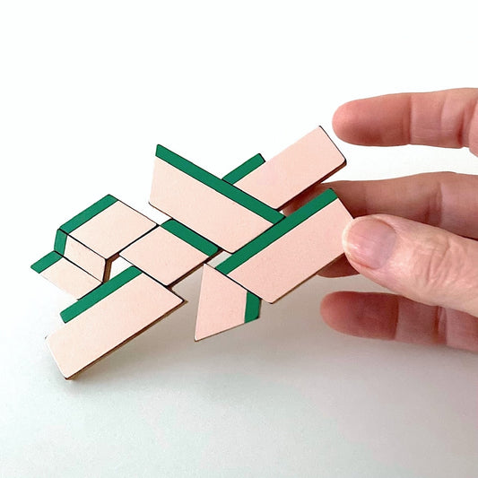 Broche XL en bois et formica rose soulignée de bandes vertes, jouant sur les lignes et la perspective. Créée à la main par l’artiste belge Karen Vanmol. Détails du bijou présenté par une main.