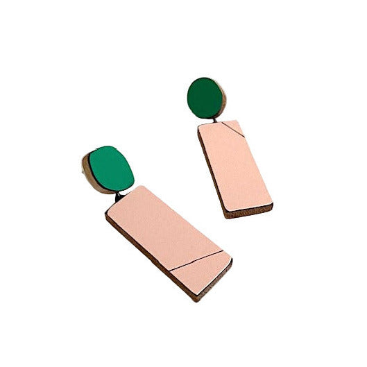 Boucles d’oreilles graphiques en bois et formica composées d'un rectangles rose pâle suspendu à un ovale vert sur l'oreille.