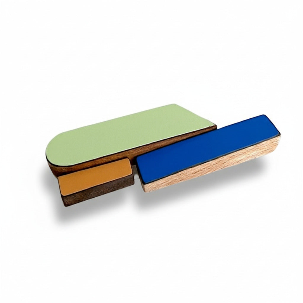 Broche contemporaine allongée en bois et formica bleu, vert et ocre. Pièce unique réalisée à la main par l’artiste belge Karen, au design graphique et coloré. 