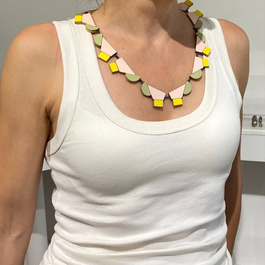 Ce collier design réunit 12 éléments géométriques en bois et formica rose poudré, vert, pastel et  jaune clair reliés par des attaches en argent patiné noir et fermés par un fermoir crochet. Bijou contemporain fait à la main par l'artiste belge Karen Vanmol. Il offre une présence graphique et élégante sur un décolleté.