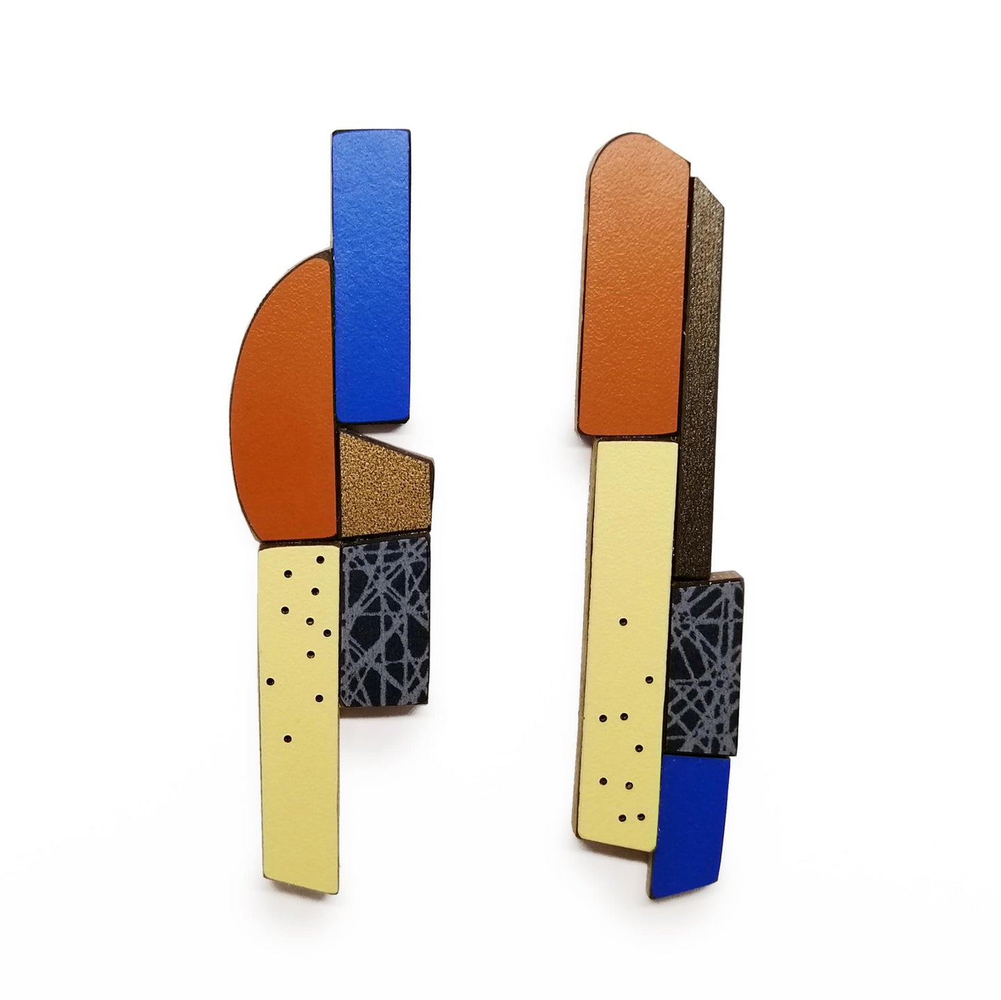 Ces boucles d’oreilles longues et asymétriques associent des éléments géométriques en bois et formica assemblés comme un jeu de construction. Pièces uniques, réalisées à la main par l’artiste belge Karen, elles mêlent des couleurs multiples : bleu foncé, marron chaud, jaune pâle, bronze métallisé aux finitions mates, avec motifs texturés. 