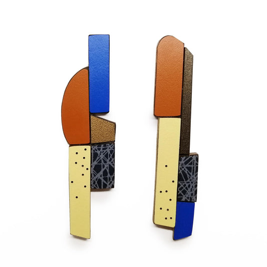 Ces boucles d’oreilles longues et asymétriques associent des éléments géométriques en bois et formica assemblés comme un jeu de construction. Pièces uniques, réalisées à la main par l’artiste belge Karen, elles mêlent des couleurs multiples : bleu foncé, marron chaud, jaune pâle, bronze métallisé aux finitions mates, avec motifs texturés. 