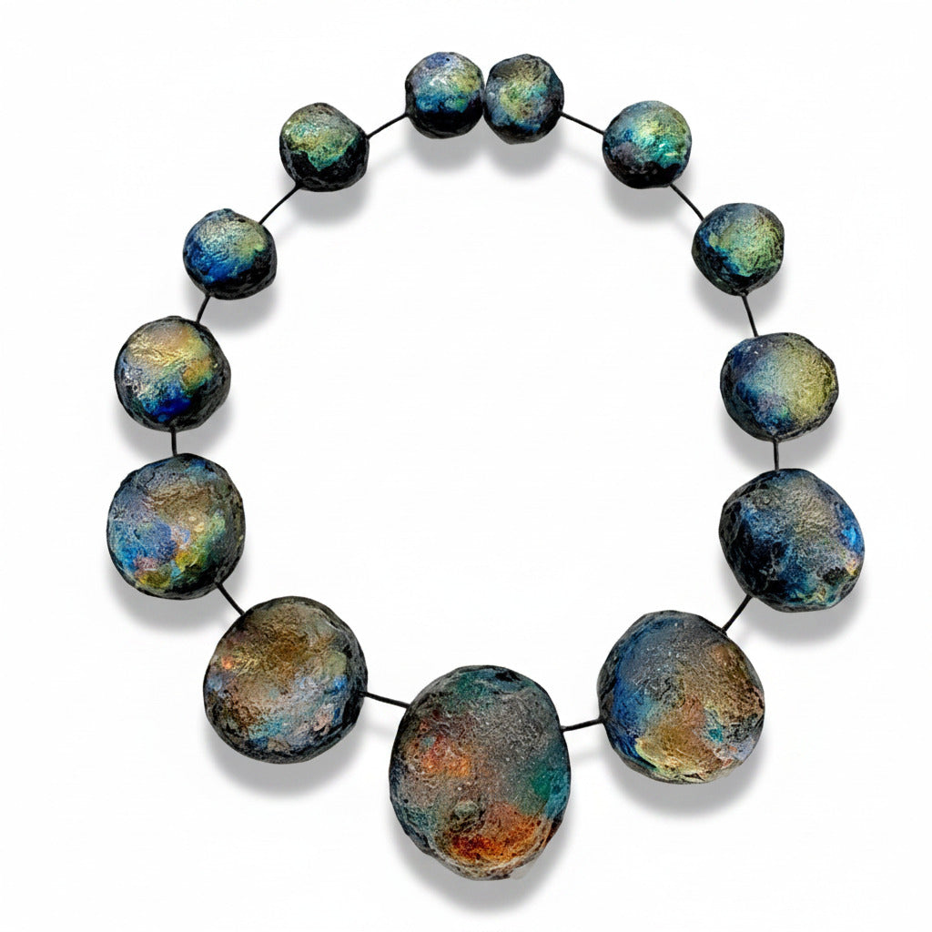Grand collier sculpturale composé 13 sphères de tailles différentes en résine texturée et irisée, dans des nuances de noir bleuté, vert et reflets changeants. Les éléments sont assemblés sur un câble fin noir et résistant. Fermeture aimantée. 