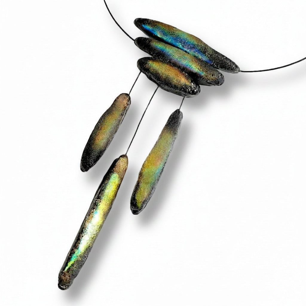 Détail du collier graphique, léger, inspiré des stalactites naturelles. Trois élément de résine texturée et irisée sont présentés horizontalement sur  un câble fin noir, ce bijou se prolonge  par trois éléments suspendus de différentes  longueurs. 