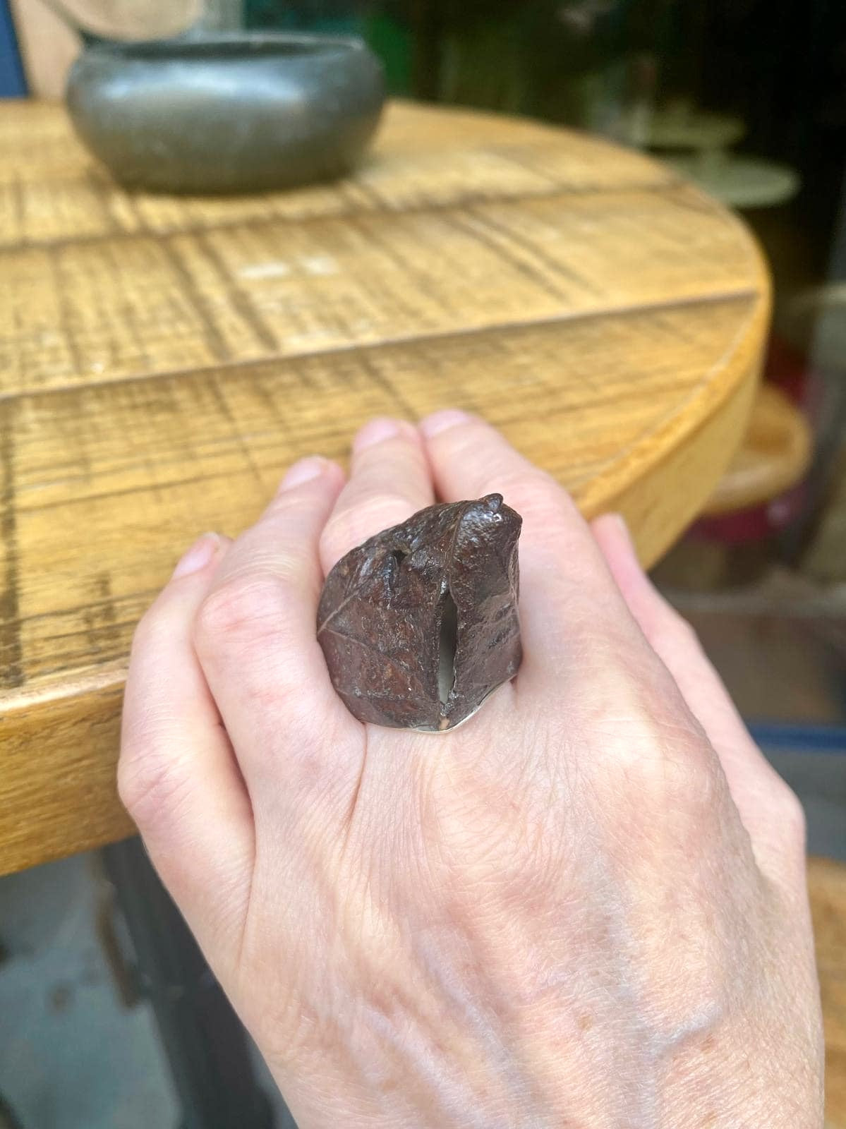 Bague haute en fer rouillé et patiné, née de moulages de feuilles d'arbres. Elle est composée d'un anneau large en argent en force de douille offrant confort et maintien. Bague portée, main posée sur une table en bois.