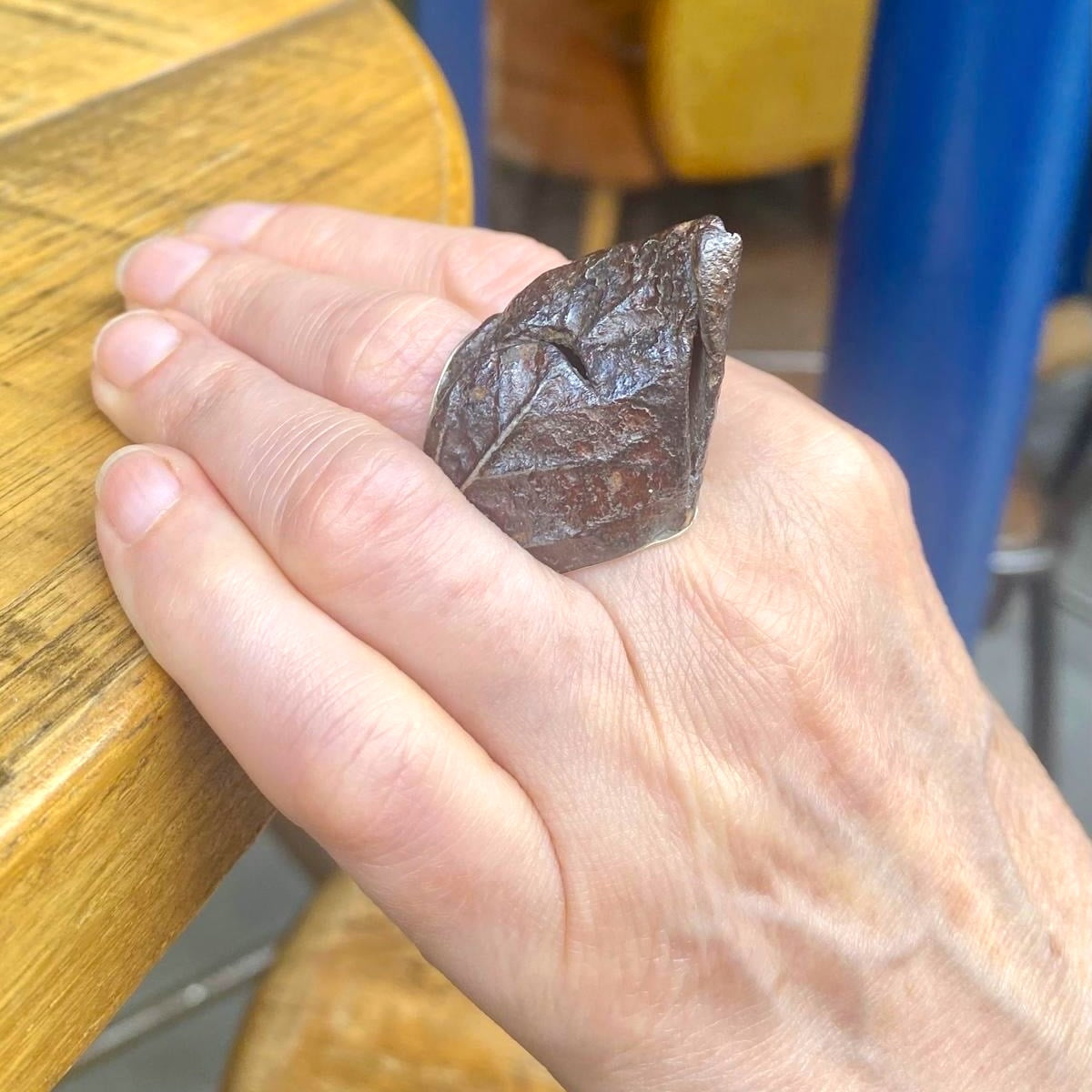 Bague haute en fer rouillé et patiné, née de moulages de feuilles d'arbres. Elle composée d'un anneau large en argent en force de douille offrant confort et maintien. Bague portée, décor table en bois.