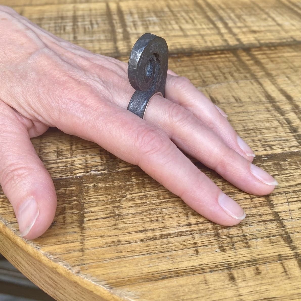Bague haute en spirale, inspirée par la forme d'une feuille de fougère enroulée. Forgée dans du fer pur, cette forme aérienne repose sur un anneau large en argent. Bijou porté, main posée sur table en bois.