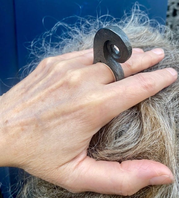 Bague haute en spirale, inspirée par la forme d'une feuille de fougère enroulée. Forgée dans du fer pur, cette forme aérienne repose sur un anneau large en argent. Bijou porté, main dans les cheveux.