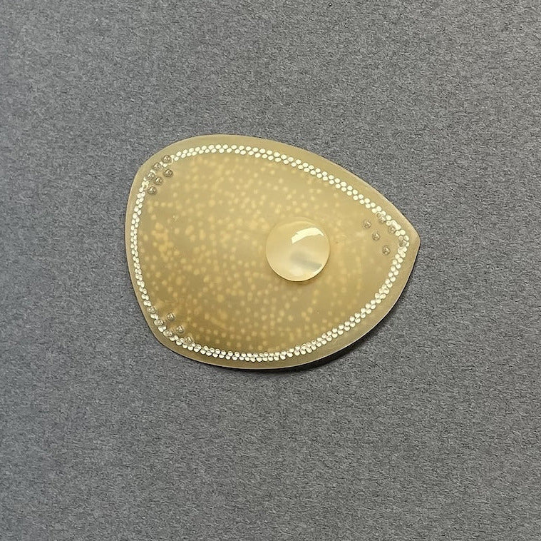 Broche en corne dorée, ornée d’une délicate frise composée de deux rangées de petits points blancs peints à la main. Une pastille de nacre ivoire apporte lumière et éclat. Envers du bijou texturé et peint.